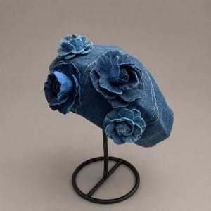 May include: A blue denim beret featuring four fabric flower appliqués made from the same denim material. The hat is constructed with visible white stitching along the seams and is displayed on a black metal stand to show its rounded shape.