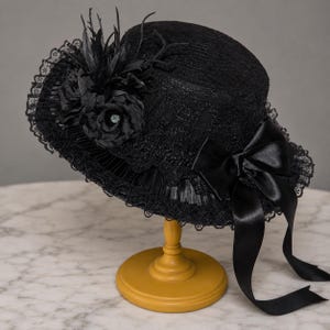 May include: A black hat featuring intricate lace detailing, a pleated brim, and a satin ribbon bow. The hat is adorned with a cluster of black fabric flowers, feathers, and thin twig-like accents. The lace trim edges the brim, adding texture to the design.