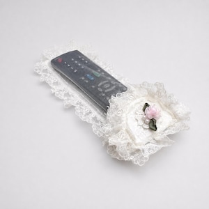 May include: A black remote control encased in a protective clear plastic cover, surrounded by a decorative white lace border. The lace features a ruffled edge and is accented with a small, pale pink fabric flower and tiny white faux pearls.