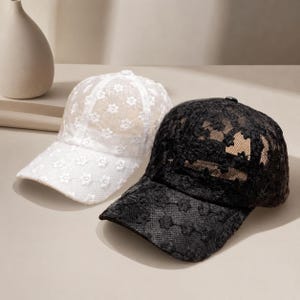 Puede incluir: Dos gorras de béisbol, una blanca y otra negra, ambas con un intrincado patrón de encaje floral superpuesto. Cada gorra tiene una corona estructurada, una visera curva y un pequeño botón en la parte superior central. El encaje es semitransparente.