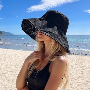 Op de afbeelding: Een zwarte bucket hat met een brede rand en een ingewikkeld bloemenkantpatroon. De hoed heeft een gestructureerde kroon en een naar beneden lopende rand die schaduw biedt. De kanten stof heeft een delicate, semi-transparante textuur.