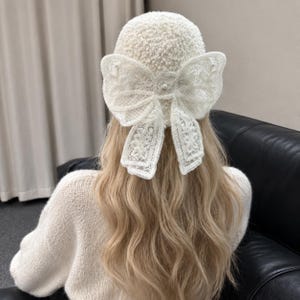 Peut inclure: Un bonnet texturé de couleur crème orné d'un grand nœud décoratif en dentelle blanche fixé à l'arrière. Le nœud est confectionné dans un tissu en dentelle transparente à motifs floraux et agrémenté d'une petite perle centrale.