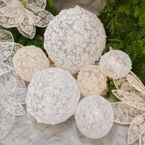 Puede incluir: Una colección de adornos esféricos y piezas decorativas con forma de flor cubiertas de tela de encaje blanco con intrincados patrones florales. El conjunto incluye varias esferas de distintos tamaños y varios recortes de encaje planos.