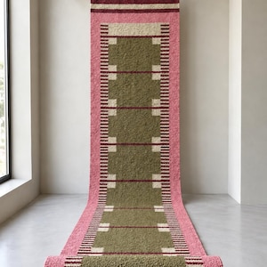 May include: A long, rectangular runner rug featuring a textured olive green center with a repeating geometric pattern of cream squares and dark red horizontal lines, bordered by wide pink edges and finished with a fringed top edge.