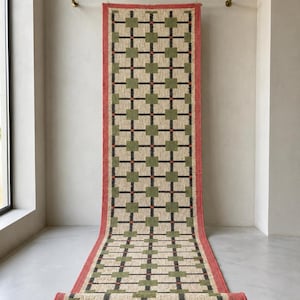 May include: A long rectangular runner rug featuring a repeating grid pattern of green squares connected by black lines on a beige background, framed by a solid red border. The text JuteEmpire is visible in the upper left corner.