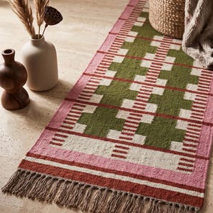 May include: A rectangular woven runner rug featuring a geometric pattern in olive green, cream, and pink, with a dark red border and fringed edges. The rug displays a repeating block design with horizontal stripes and a textured, knotted surface.