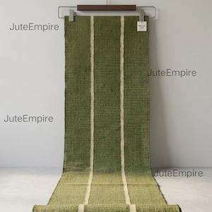 May include: A rectangular olive green runner rug with two vertical cream-colored stripes running down the length. The rug is suspended from a wooden hanger with metal clips. The text JuteEmpire appears multiple times, and a small tag reads Natural Jute.