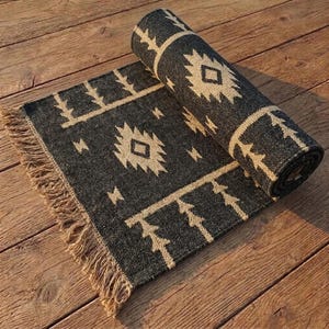 May include: A rectangular area rug featuring a dark charcoal gray base with a contrasting cream-colored geometric pattern. The design includes diamond shapes and linear motifs, finished with long, natural-toned fringe along one edge.