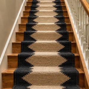 May include: A stair runner featuring a textured, woven design with a central tan diamond pattern bordered by solid black edges. The runner is installed on a wooden staircase, covering the treads and risers in a repeating geometric sequence.
