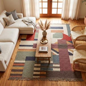 May include: A rectangular area rug featuring a geometric pattern in shades of red, blue, green, and brown with fringe on the ends. The rug is placed on a wooden floor beneath a wooden coffee table, a white sectional sofa, and two rattan chairs.