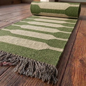 May include: A woven runner rug featuring a repeating geometric pattern of olive green and cream-colored shapes. The rug is partially rolled at one end and finished with a fringe of loose, textured gray fibers along the bottom edge.