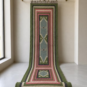 May include: A long rectangular runner rug featuring a geometric pattern with concentric borders in olive green, dusty pink, cream, and charcoal gray. The center displays a light blue diamond motif. The rug has fringed ends and the text JuteEmpire.