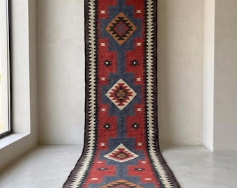 May include: A long runner rug featuring a geometric pattern in red, blue, black and cream. The rug is suspended from a gold-toned metal rod and features a rolled end at the bottom. The text JuteEmpire is visible in the top left and bottom right.