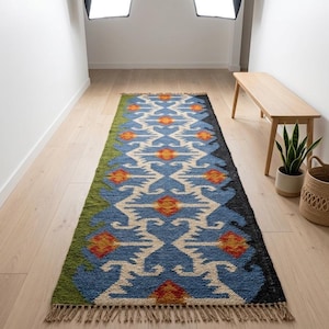 May include: A long rectangular runner rug featuring a repeating symmetrical pattern in blue, cream, orange, and green. The rug has a textured pile, a fringed edge on the short side, and a solid green border on one long side and black on the other.