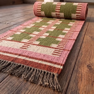 May include: A woven runner rug featuring a geometric grid pattern in olive green, cream, and terracotta red. The rug has a rolled end and a flat end finished with a fringe of brown tassels. The textile displays a textured, multi-colored weave.