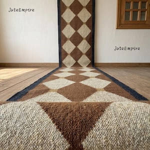 May include: A long runner rug featuring a repeating diamond pattern in alternating shades of brown and cream. The rug has dark blue borders along the sides and is displayed hanging from a metal rod, with the bottom portion rolled onto the floor.