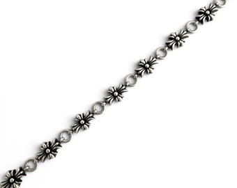 Puede incluir: Una pulsera de cadena plateada con un patrón repetitivo de pequeños dijes ornamentados en forma de cruz unidos por anillos metálicos circulares. La superficie metálica tiene un acabado pulido con áreas oscurecidas en relieve.