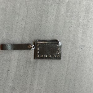 Könnte beinhalten: Eine dunkelgraue, metallisch glänzende rechteckige Geldbörse mit strukturierter Oberfläche, einem Reißverschluss an der Oberseite und einer passenden Handschlaufe, die an einem silbernen Metallring befestigt ist. Die Vorderseite ist mit Nieten verziert.