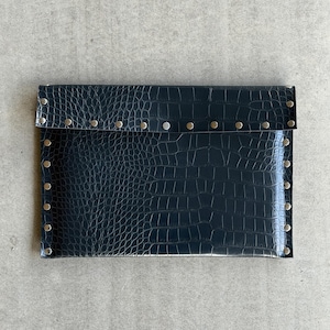 May include: A rectangular black clutch bag featuring a textured crocodile skin pattern. The edges and the fold-over flap are secured with evenly spaced silver-toned metal studs that run along the perimeter and the horizontal closure line.