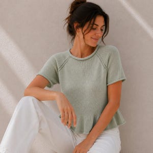 May include: A sage green short-sleeved knit top featuring a round neckline, raglan sleeves with delicate openwork detailing along the seams, and a subtle flared peplum hem. The garment is paired with plain white trousers.