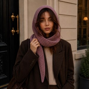 Op de afbeelding: Een mauve gebreide capuchonsjaal met een geribbelde textuur die het hoofd bedekt en over de borst valt. Het accessoire wordt gedragen over een beige top en een donkerbruine blazer, met een puntig uiteinde en een consistent breipatroon.