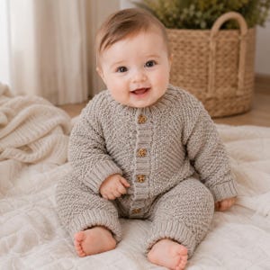 Op de afbeelding: Een lichtbeige gebreide romper met lange mouwen, voorzien van een gestructureerd breipatroon, een ronde halslijn en een knoopsluiting aan de voorzijde met vier kleine, bruin gespikkelde knoopjes. Het kledingstuk heeft geribbelde boorden bij de polsen en enkels.