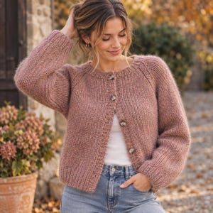 Könnte beinhalten: Eine altrosa, langärmelige Strickjacke aus grobem Strick mit Rundhalsausschnitt und vier großen, gesprenkelten braunen Knöpfen auf der Vorderseite. Das Kleidungsstück hat gerippte Bündchen an Ärmeln und Saum sowie eine strukturierte Optik.