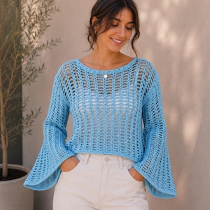 May include: A light blue long-sleeve crochet sweater featuring an open-knit pattern, a wide boat neckline, and flared bell sleeves. The garment is worn over a simple white camisole and paired with light-colored trousers, creating a layered look.