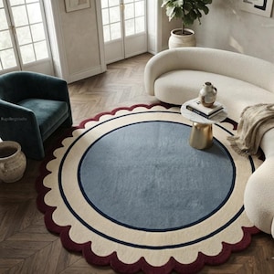 May include: A large round area rug featuring a central dusty blue circle, a wide cream-colored border, and a dark navy blue ring accent. The outer edge of the rug is finished with a scalloped design in a deep burgundy red color.