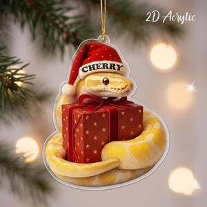 May include: A 2D acrylic Christmas ornament featuring a yellow snake wearing a red Santa hat with the name CHERRY printed on the brim. The snake is coiled around a red gift box decorated with gold stars and a red ribbon bow, hanging by a gold cord.