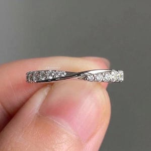 Könnte beinhalten: Ein silberfarbener Metallring mit gedrehtem Banddesign. Die Oberfläche des Rings ist mit einer durchgehenden Reihe kleiner, klarer, rund geschliffener Steine besetzt, die funkeln. Der Text "joliediamond" ist in kleinen weißen Buchstaben auf dem Ring.