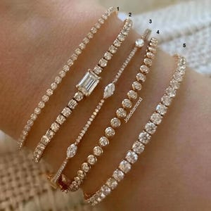 Può includere: Cinque bracciali tennis color oro numerati da uno a cinque. Il bracciale uno è una sottile catena di pietre rotonde. Il bracciale due presenta una pietra centrale taglio smeraldo. Il bracciale tre ha piccole pietre marquise. Il bracciale quattro ha pietre rotonde incastonate. Il bracciale cinque è un classico bracciale tennis con pietre rotonde.
