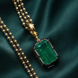 May include: A rectangular emerald pendant set in a dark metal frame, suspended from a gold-toned ball chain. The pendant features a large, faceted green gemstone held by prongs, with a polished gold-toned bail connecting it to the chain.