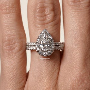 Peut inclure: Une bague en argent avec une pierre centrale en forme de poire entourée d'un halo de petites pierres rondes. L'anneau et le halo sont incrustés de petites pierres rondes supplémentaires. Le texte Joliediamond est visible sur l'anneau.