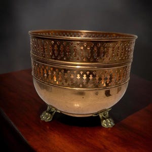 Peut inclure: Un bol en laiton poli avec une base arrondie et trois pieds en forme de griffe. La partie supérieure du bol présente une bande décorative avec un motif répétitif de découpes verticales, créant un effet de galerie perforée sur le bord.
