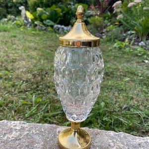 Op de afbeelding: Een decoratieve pot van helder glas met een textuur van herhalende ovalen op het lichaam. De pot is voorzien van een gepolijste goudkleurige metalen voet en een bijpassend goudkleurig metalen deksel met een puntige knop aan de bovenzijde.