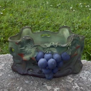 Op de afbeelding: Een keramische plantenbak met een sculpturaal reliëf van paarse druiven en groene bladeren aan de voorzijde. De pot heeft een matte afwerking met een gedempte groene basiskleur, een open bovenkant met geschulpte randen en decoratieve uitsparingen.