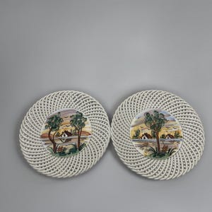 May include: Two round ceramic plates featuring a white lattice-style woven border and a central hand-painted scene depicting a landscape with trees, a small house, and a body of water in shades of green, brown, yellow, and blue against a white background.