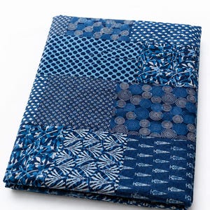 May include: A folded textile featuring a patchwork design of various blue and white patterns. The fabric displays geometric shapes, including circles, dots, leaf motifs, and linear designs, all rendered in shades of indigo and white on a cotton-like surface.