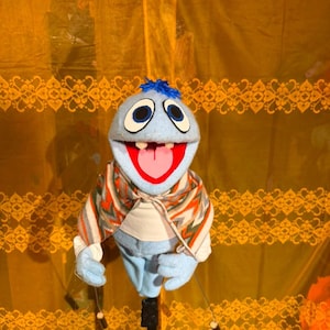 May include: A light blue hand puppet with a tuft of blue hair, large white eyes with black pupils, and an open mouth showing a pink tongue and two white teeth. The puppet wears a white shirt and a patterned shawl with orange, grey, and green stripes.
