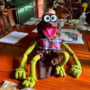 May include: A brown puppet with large white eyes and black pupils, wearing a red and blue plaid vest with a single brown button. The puppet has long, light green arms and hands, a wide open mouth with a pink tongue, and a brown textured body.