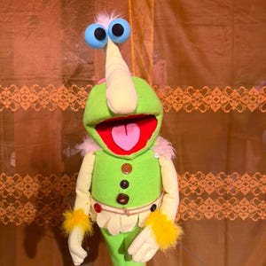 May include: A green puppet with a long, pointed nose, large blue eyes, and a wide open mouth with a pink tongue. The puppet features yellow faux fur cuffs, a cream-colored vest with three brown buttons, and a small white button on the shoulder.