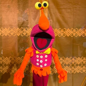 May include: A plush puppet featuring a bright magenta torso with six white buttons, a yellow bow tie, and orange arms with ruffled cuffs. The head has a large orange nose, two yellow eyes on stalks, and an open mouth with a red heart-shaped tongue.
