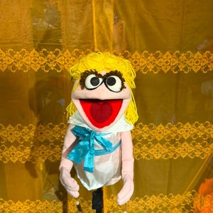 May include: A hand puppet with light pink skin, bright yellow yarn hair, and large black-rimmed eyes. The puppet features a wide open red mouth and wears a white sleeveless dress with a shiny blue ribbon tied in a bow at the waist.