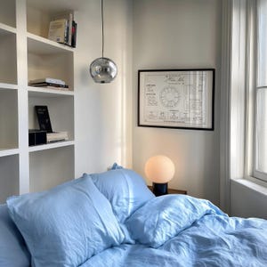 May include: A light blue bedding set with pillows and a duvet cover, positioned in a room with white built-in shelves containing books, a spherical chrome pendant light, a round frosted glass table lamp, and a framed astrological chart.