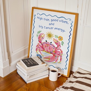 May include: A framed art print featuring a pink crab illustration with yellow flowers and the text "High tide, good vibes, and big Cancer energy." The print is bordered by a blue wavy line and housed in a light wood frame, leaning against a wall.