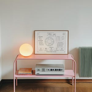 May include: A rectangular framed astrological chart with black text and graphics on a white background, featuring a central circular zodiac wheel, numerology data, and the name Melissa Niles, displayed on a pink metal console table.