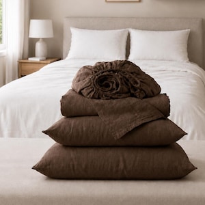 Pode incluir: Uma pilha de roupa de cama castanho-escura composta por um lençol com elástico, um lençol plano e duas fronhas retangulares. Os artigos estão dobrados e empilhados, exibindo uma superfície de tecido texturizado num tom castanho terroso profundo.