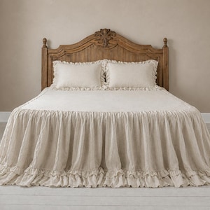 May include: A beige bedspread with a double-layered ruffled skirt and two matching rectangular pillows. The bedding is set against a tall, arched wooden headboard with vertical paneling and a carved decorative detail at the centre top.