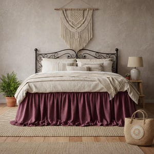 May include: A bed with a black metal frame, a cream-coloured duvet, and a deep plum-coloured bed skirt. The bed is dressed with several cream pillows and a throw blanket. Above the headboard hangs a large, cream-coloured macrame wall hanging.